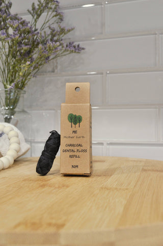 A spool of dental floss placed in front of a cardboard packaging labeled 'Me Mother Earth - Eco Floss- Refill ONLY - One Refill' on a wooden surface with a brick wall in the background.
