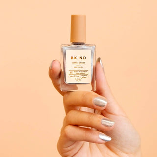 A hand holding a bottle of BKIND nail polish in a clear color, with a label that reads 'Vegan. 77% plant-based. 21-FREE.'