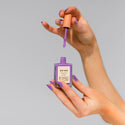 A hand holding a bottle of BKIND nail polish in a purple color, with a application brush on top.