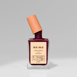 A bottle of BKIND nail polish in a dark purple shade with a glossy finish, featuring a brush applicator and labeled as vegan and 21-free.