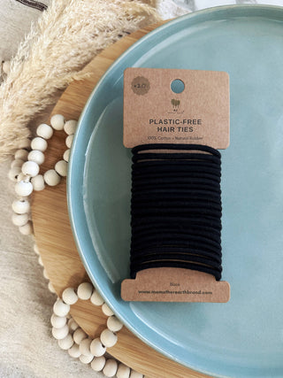 A bundle of black plastic-free hair ties placed on a blue plate, with a cardboard packaging label indicating the product is plastic-free and made with natural materials.